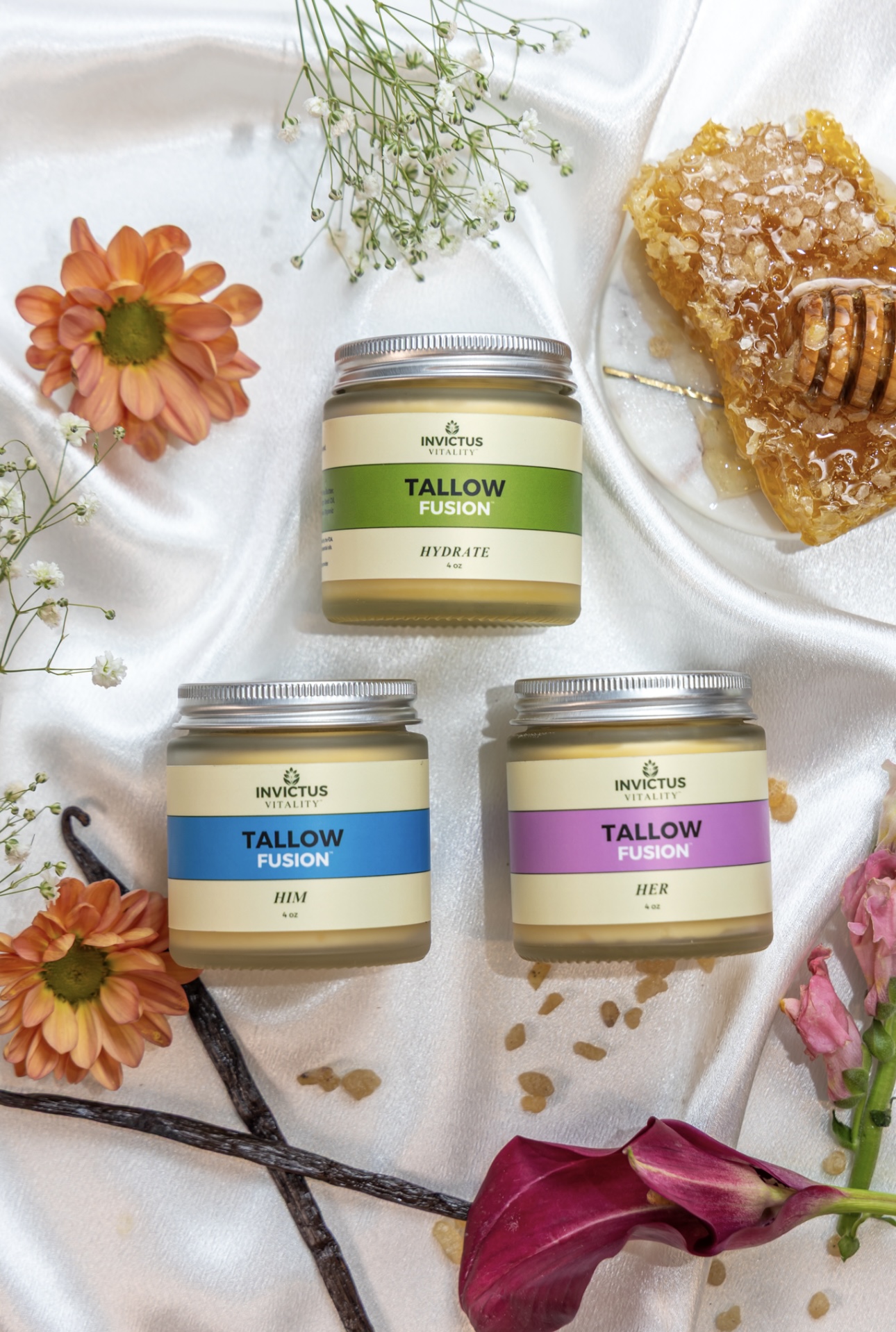 Three original Invictus Vitality Tallow Fusion jars on white fabric with flowers, honeycomb, and vanilla beans